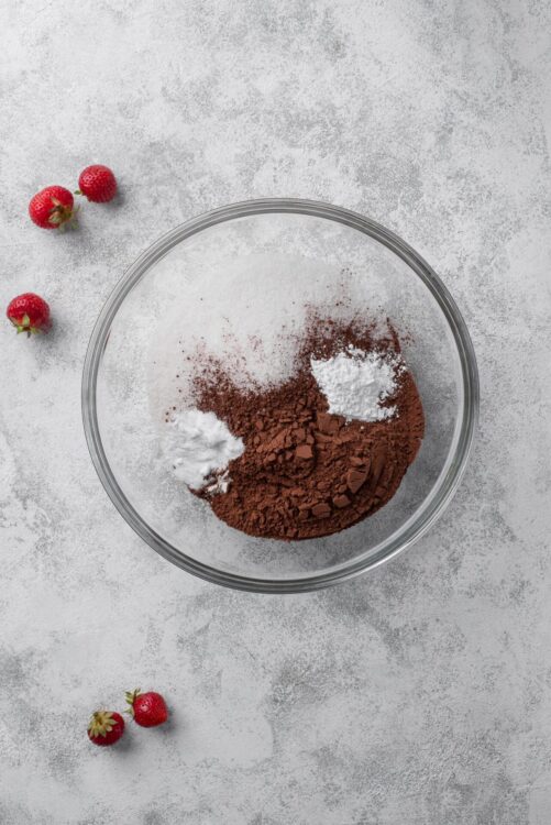 Rich cocoa powder, flour, and sugar in a glass bowl for baking, with fresh strawberries on a textured surface.