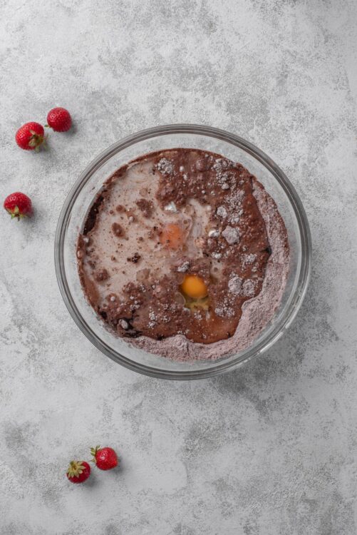 Eggs and cocoa powder in a mixing bowl for baking, on a textured surface with strawberries nearby.