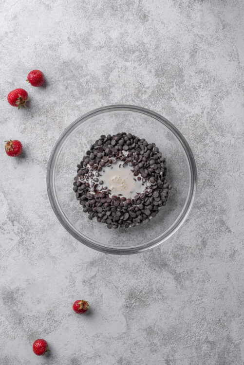Fresh chocolate chips and milk in a glass bowl surrounded by strawberries on a textured gray surface.