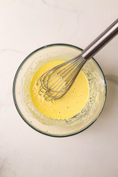 Light yellow homemade cake batter in a glass mixing bowl with a metal whisk on a white surface.