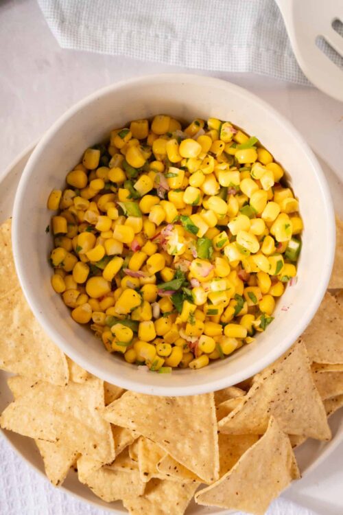 Corn salsa with chopped green onions and red onions in a white bowl, served with tortilla chips on a white plate, fresh and flavorful snacks for parties, summer meals.