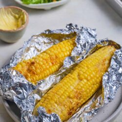 Buttered corn on the cob wrapped in aluminum foil, ready to serve with a side salad and baked dish.
