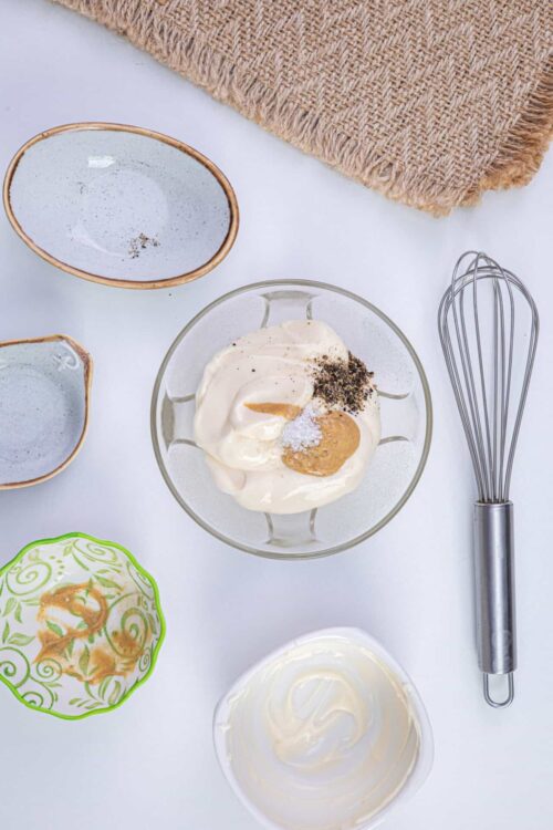 Whisk, salt, pepper, and spices for creamy dipping sauce or spread on a white table with bowls and a whisk.