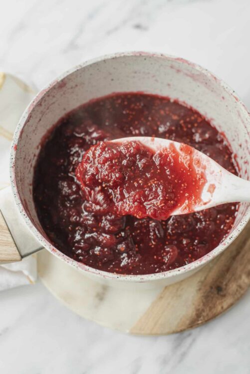 Fresh homemade berry jam in a white pot with a spatula on a marble surface.