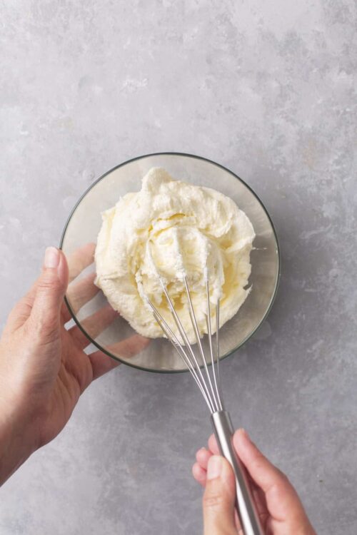 Cream cheese frosting in a glass bowl with a handheld mixer, ready to spread or pipe for baked goods.