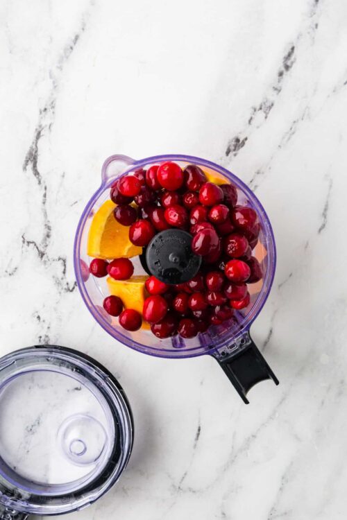 Fresh fruit in a blender, including red currants and peach slices, on a white marble surface.