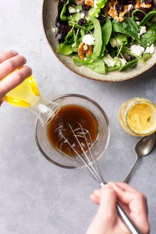 Balsamic vinaigrette being whisked over mixed greens salad with walnuts and feta cheese.