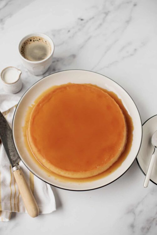 Sweet caramel flan dessert in a white bowl on marble surface, topped with shiny caramel sauce.