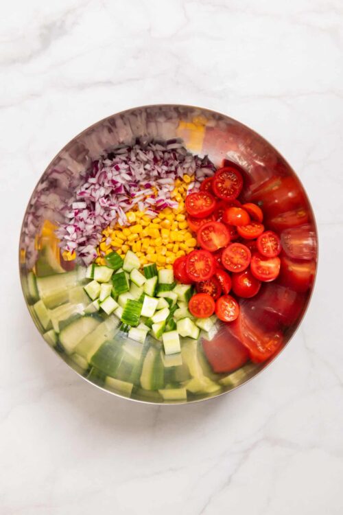 Diced red onions, corn, cherry tomatoes, and cucumber in a stainless steel bowl on white marble surface.