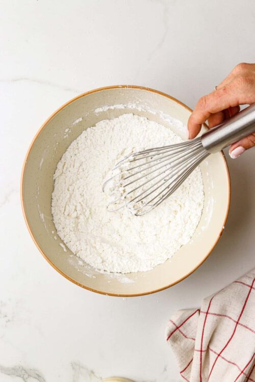 Flour being mixed in a beige bowl with a metal whisk, illustrating baking process.