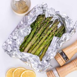 Baked asparagus wrapped in foil with lemon slices and seasoning on a white background.
