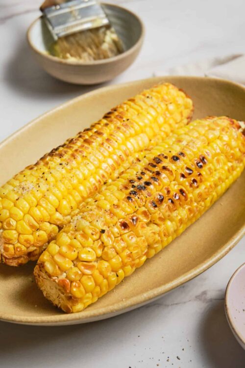 Golden grilled corn on the cob with charred kernels, served on a beige platter for a baked Bree recipe.