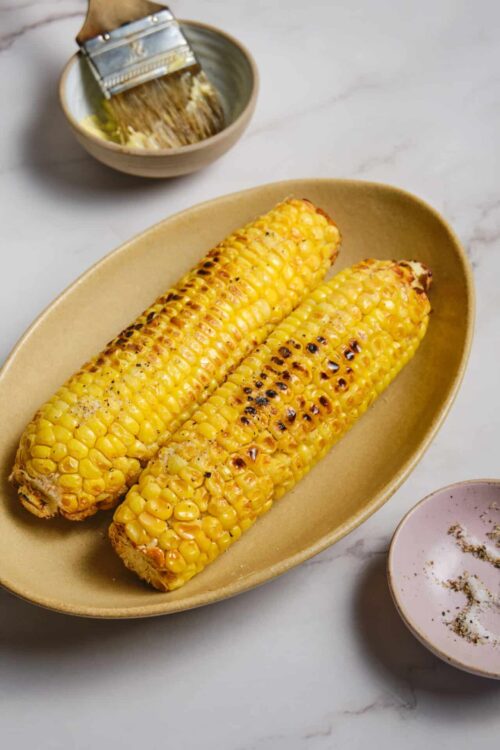 Buttered grilled corn on a beige platter with seasoning and char marks.
