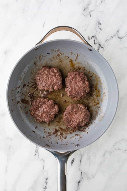 Juicy homemade burger patties cooking in a skillet with browned edges and rendered fat.