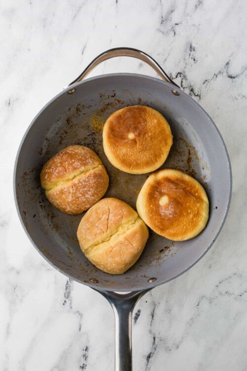 Buttered english muffins in a cast iron pan, toasted golden brown, perfect for breakfast recipes, baked bread, or English muffin sandwiches.
