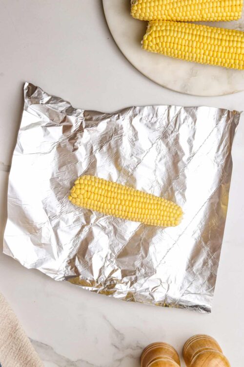 Fresh yellow corn on aluminum foil ready for cooking, with additional corn cobs and cooking essentials.