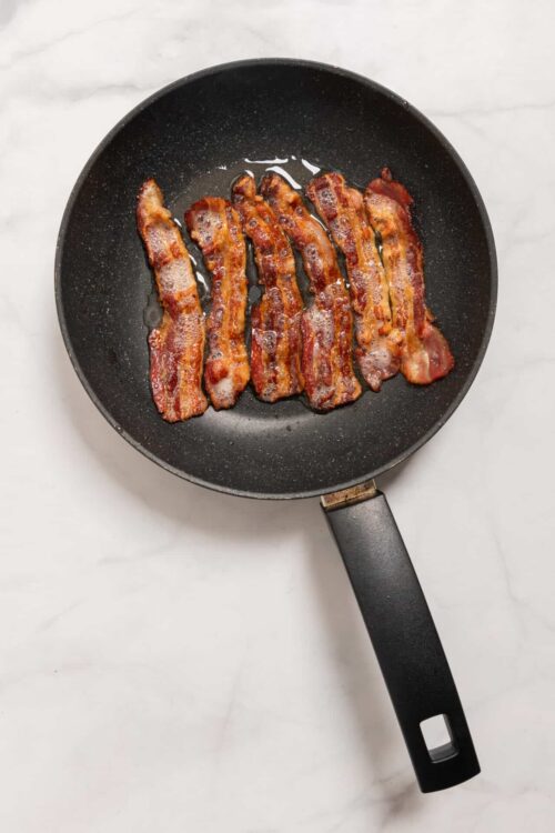 Crispy bacon strips cooking in a black frying pan on a white marble countertop.