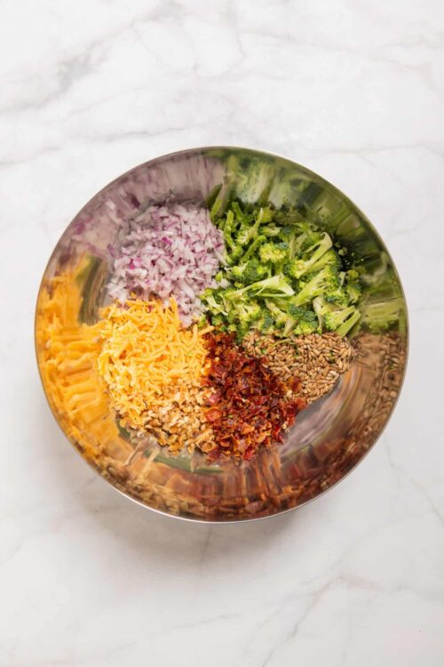 Chopped red onions, broccoli florets, shredded cheddar cheese, sun dried tomatoes, toasted sunflower seeds in a metal bowl.