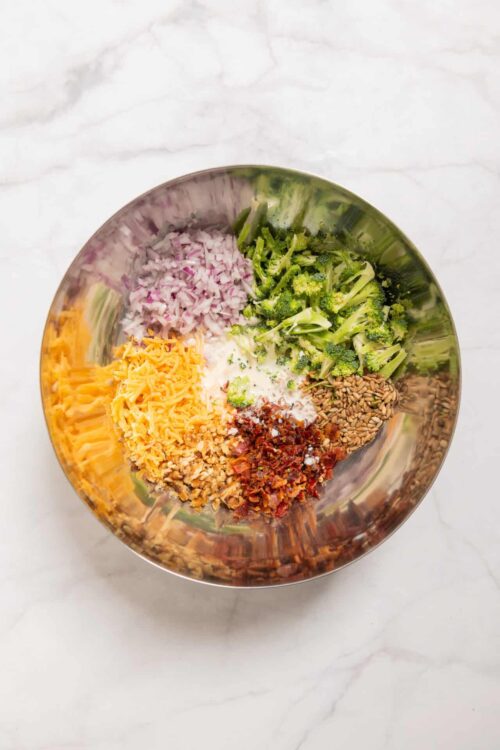 Chopped red onions, broccoli florets, shredded cheddar cheese, sunflower seeds, sun-dried tomatoes, and dressing in a stainless steel bowl.