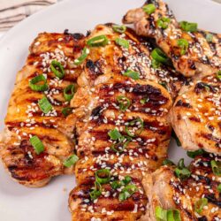 Grilled chicken breasts with sesame seeds, green onions, and barbecue glaze, on a white plate.