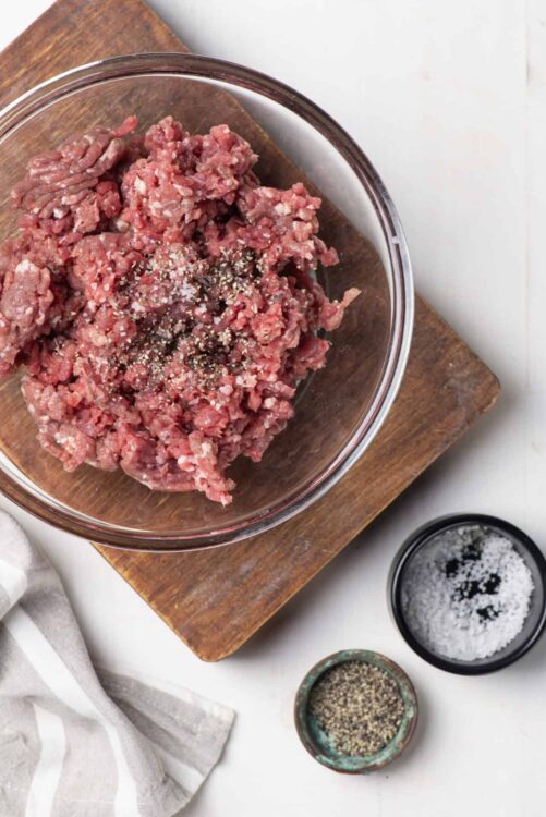 Ground beef in glass bowl with salt and pepper on white countertop.