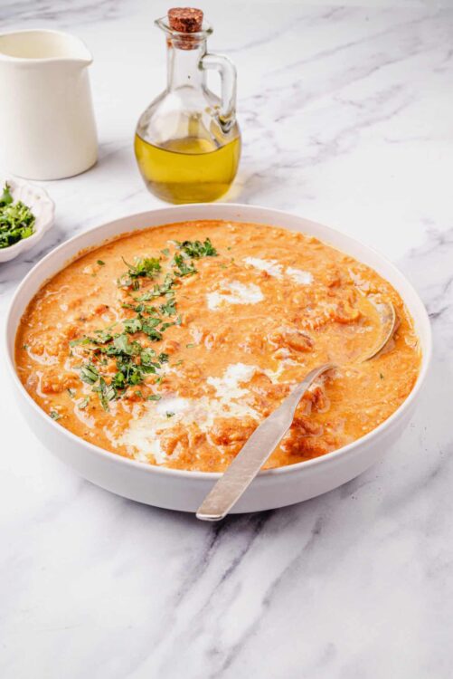 Creamy tomato soup with fresh herbs in a white bowl on marble surface, served with olive oil and garnished with cilantro.