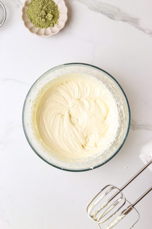Cream cheese frosting in a glass bowl, with beaters and matcha powder on a white marble surface.
