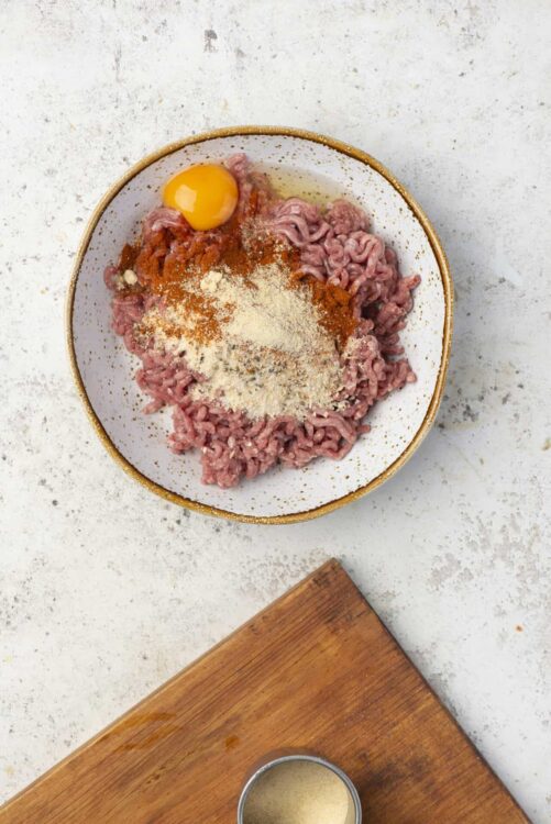 Ground beef mixture with egg, spices, and breadcrumbs in a white bowl for homemade meatloaf or burger prep.
