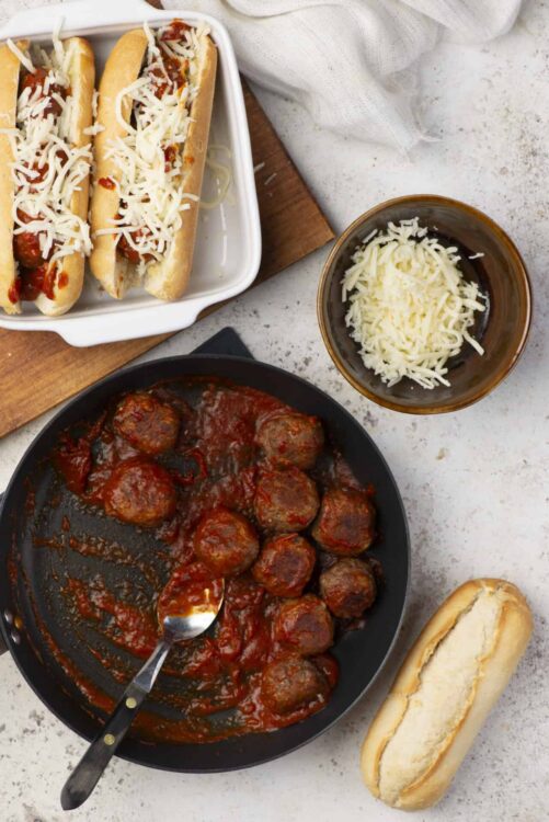 Savory Italian meatballs in marinara sauce with cheeseburger hot dogs and shredded cheese on a textured white surface.