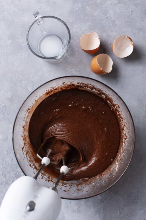 Rich chocolate cake batter being whisked in a glass mixing bowl with eggs and milk nearby.