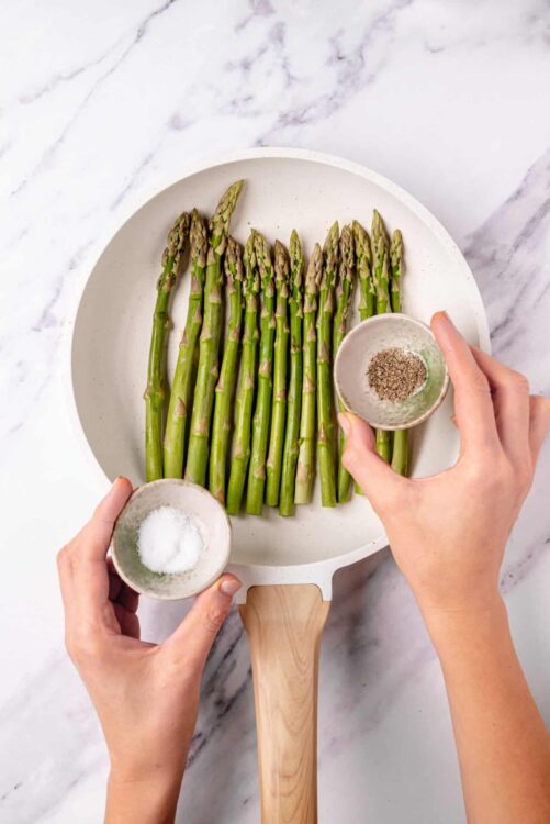 Freshly prepared asparagus spears with salt, pepper, and seasoning in a kitchen setting.