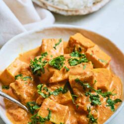 Creamy chicken curry with herbs served in a white bowl with naan bread in the background.