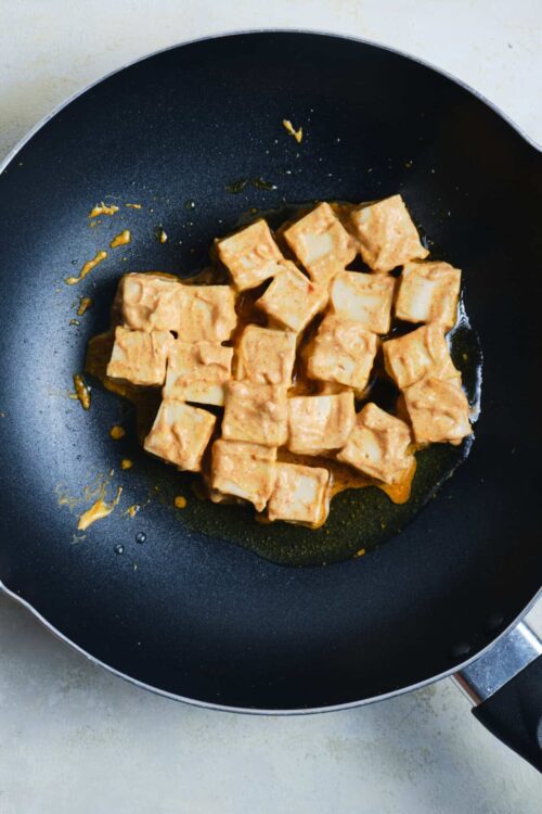 Creamy tofu cubes cooking in a black skillet with olive oil, on a light countertop.