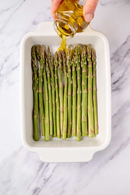 Fresh asparagus being drizzled with olive oil in a white baking dish, ready for roasting or baking.