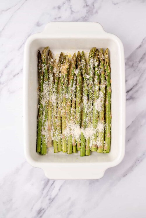 Roasted asparagus with grated Parmesan in a white baking dish, on a marble surface.