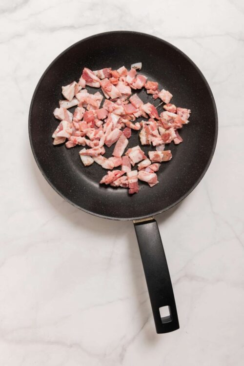 Crispy chopped bacon cooking in a black skillet on a white marble countertop.