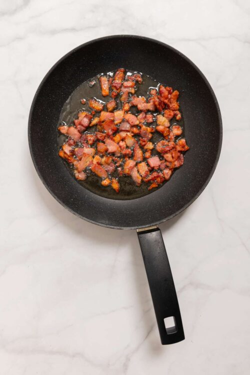 Crispy bacon bits cooking in a black skillet on a white marble surface.