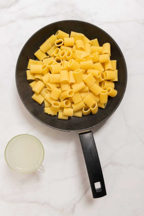 Cooked rigatoni pasta in a black skillet with a glass of water nearby.