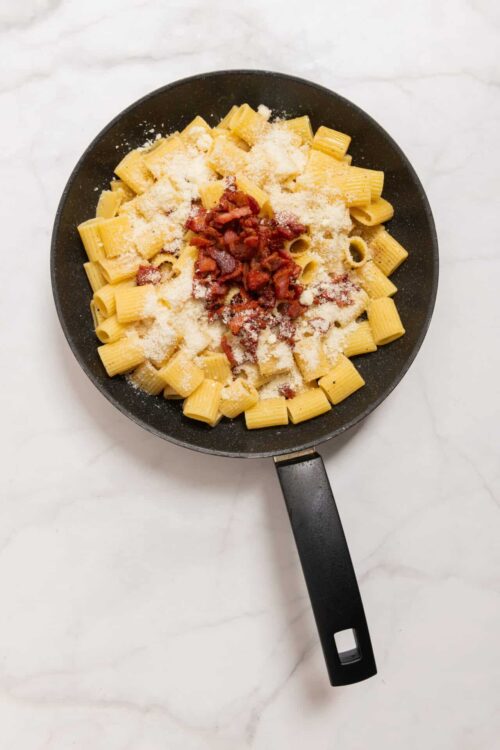 Creamy pasta with bacon and grated cheese in a black skillet, ready to bake for a cheesy pasta dish.