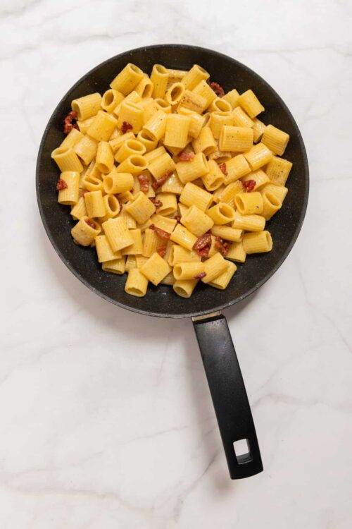 Creamy pasta with bacon in a black skillet on white marble surface.