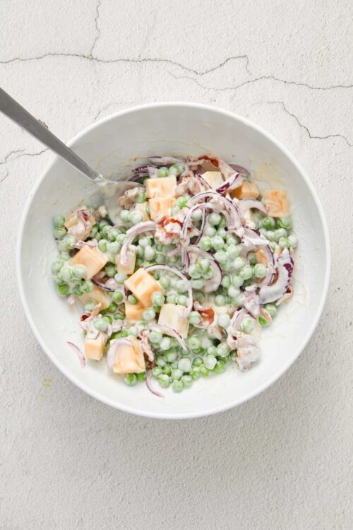 Creamy pea and cheese salad with onion and bacon in a white bowl.