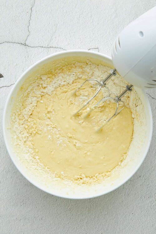 Creamy cake batter being mixed with electric hand mixer in a white bowl, baking ingredients for homemade cake.