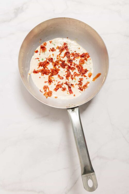 Crispy bacon bits in creamy white sauce in stainless steel skillet.