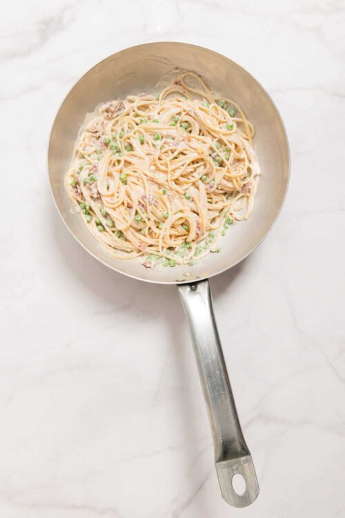 Creamy salmon pasta with peas in a stainless steel skillet on white marble surface.