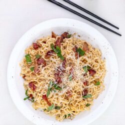 Creamy bacon and ramen noodle dish on a white plate with shredded cheese and green onions.
