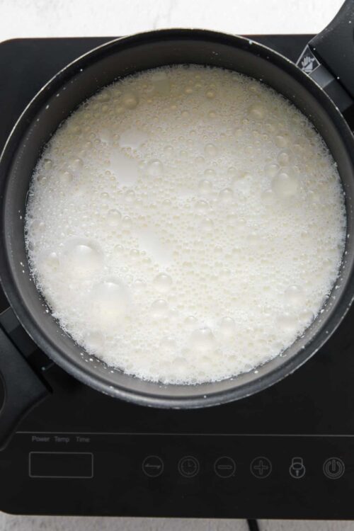 Creamy milk being heated in a black saucepan on an induction stove.