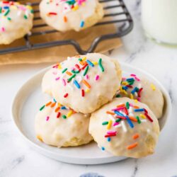 Colorful sprinkle coated sugar cookies on white plate with a cooling rack in background.