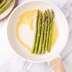 Fresh green asparagus being cooked in a white skillet with butter.
