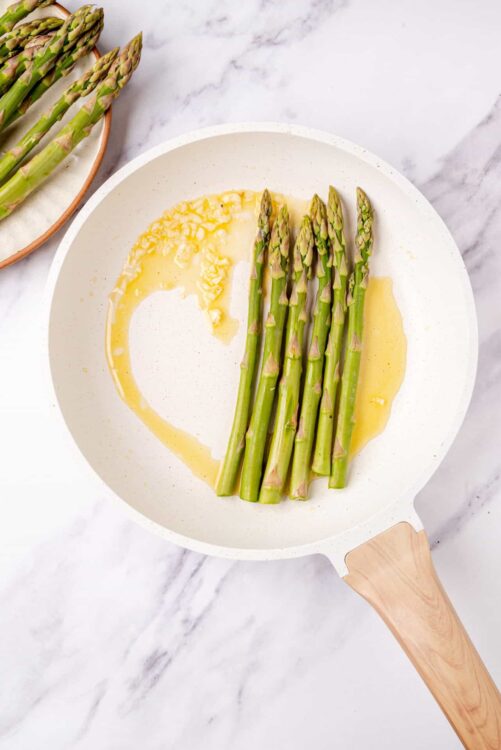 Fresh green asparagus being cooked in a white skillet with butter.