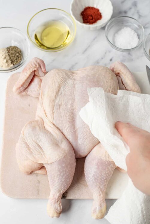 Raw whole chicken on cutting board with seasoning ingredients, olive oil, and paper towels.